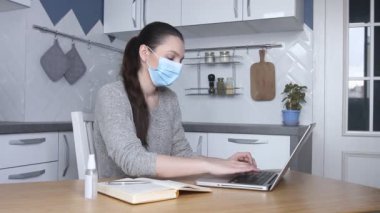 Tıbbi maske takan genç bir kadın dizüstü bilgisayarında e-posta yazarak serbest çalışıyor. Ev mutfağında çalışmak için bilgisayar kullanan bir kadın, uzaktan internet işi.