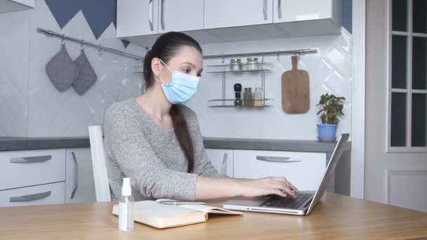 年轻的女自由职业者戴着医疗面具 在家工作 在笔记本电脑上输入电子邮件 使用电脑上网的女性在家厨房 远程上网工作 — 图库视频影像