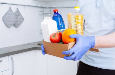Koruyucu tıbbi eldivenlerdeki kurye, modern mutfak arka planında karantina için taze çiğ sebze ve yiyecek stokları içeren bir kutu teslim ediyor. Organik gıda ve ürün teslimatı