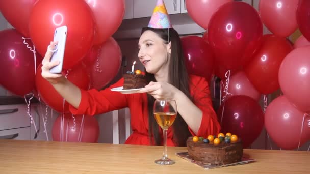 誕生日の帽子の若い女性ブロガーは ろうそくとケーキの作品を保持携帯電話に話しています 幸せな誕生日の女性だけで自宅でオンライン会議コールやライブストリーミングガジェットを使用して — ストック動画
