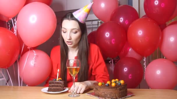 誕生日の帽子の悲しい若い女性は隔離中に台所で一人で誕生日を祝う 風船を背景にろうそくのあるケーキと一人で座って — ストック動画