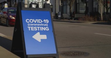 Büyük bir şehrin kaldırımında yayaları ve geçen arabaları gösteren bir tabela var. Yerel COVID-19 test merkezinin olduğu yerde..