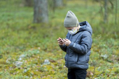 Ağızlarında koronavirüs hastalığı olan ve ormanda siyah bir telefonla oyun oynayan bir çocuk.