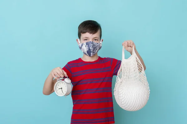 Boy in blue mask on face with mesh bag, ball and alarm clock on blue background. Coronavirus outbreak situation.
