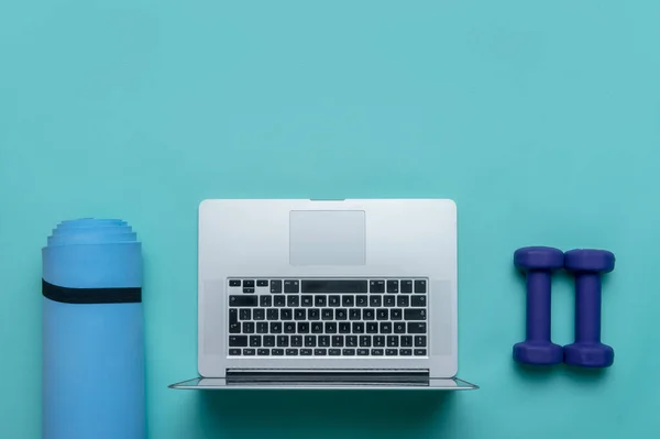 Blue dumbbells, gym mat and grey laptop on blue background. Online workout concept