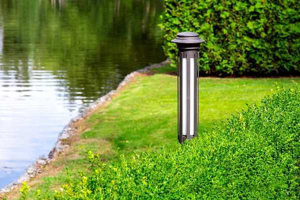 An iron garden lighting lantern mounted on a green lawn with bushes on the shore by the water pond on a sunny summer day.