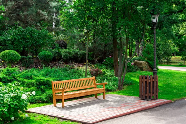 empty wooden bench with a wooden trash can and lantern in the well groomed park with trees and bushes on a summer day, 3/4 view nobody.