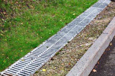 Yol kenarındaki drenaj sisteminde demir ızgarası ve yeşil çimen olan bir hendeğin arkasında..
