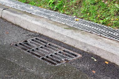 Kaldırımın arkasındaki kaldırım kenarında yağmurdan sonra asfalt bir yolda kanalizasyon ızgarası ızgara ızgarası var..