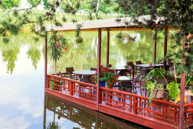 kırmızı bir iskelede yüzen bir restoran. Bir yaz günü nehir kıyısında sandalyeler ve saksılarla masaların üzerinde bir tepe örtüsü var..