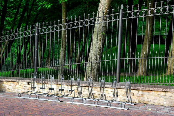 free bike parking on the sidewalk made of tiles near a black metal fence with peaks in a park with trees.