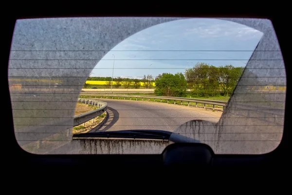 dirty car view from inside the car through the back window, rear window of a dirty car with a view of the asphalt road with turn road roadside with trees and field.