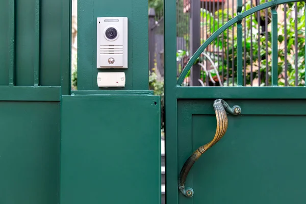 an intercom with a video camera and a microphone for voice communication with a card reader for access by a key card is installed on a green iron gate with handle, close up nobody.