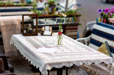 Örgü bir masa örtüsü ve cam bir şişeyle açık bir kafe masası. Çiçekli papatyalarla dolu bir vazo..