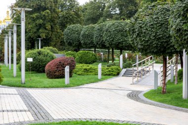 Şehir parkındaki yaprak döken ağaçların altında yeşil çimenler ve çalılarla kaplı taştan fayanslarla döşenmiş bir meydan. Yerden demir fenerler ve sokak lambaları..