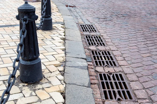 hatches of the drainage system of the road paved with paving stones with a curb and stone tiles on the sidewalk with iron columns on chains for the safety of pedestrians, nobody.