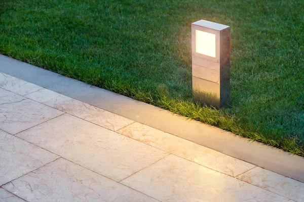 ground lamp lighting marble walkway in the evening park with a green lawn, closeup lantern illuminated marble pavement, nobody.
