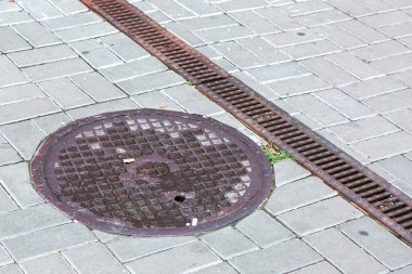 Kare taş fayanslardan yapılmış bir alanda, yağmur suyu drenaj sistemini kapatan yuvarlak bir rögar kapağı ve kanalizasyon asfalt sistemi..