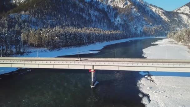 Vuelo sobre el río Katun cubierto de hielo y nieve con un puente automovilístico sobre el que viaja el transporte en las montañas Altai . — Vídeos de Stock