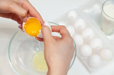 Yumurtayı parmaklarıyla kıran ve sarısını bir tabağa koyup bir düzine yumurta ve un dolu bir bardakla birlikte bir paketin arkasına döken bir kadının ellerine yakın plan.