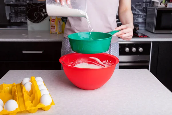 Making dough for Easter cake at home in the kitchen. Flour is poured out and sifted through a green sieve in female hands into a red bowl next to the ingredients - eggs in a yellow substrate.