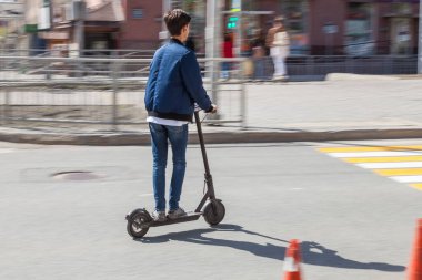 Mavi bombardıman ceketi ve elektrikli scooterlı kot pantolonlu genç bir adam - şık bir cihaz, yaya geçidinde caddeden geçiyor. Modern çevre dostu bir cihaz..