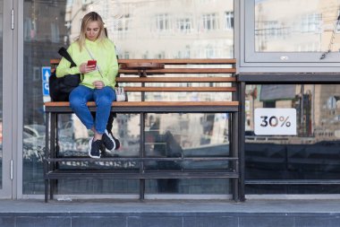 Parlak sarı kazaklı sarışın bir kız bir kahve dükkanının yanındaki tahta bir bankta oturuyor, elinde bir telefon tutuyor ve emirlerini bekliyor. Otobüs durağındaki kız..