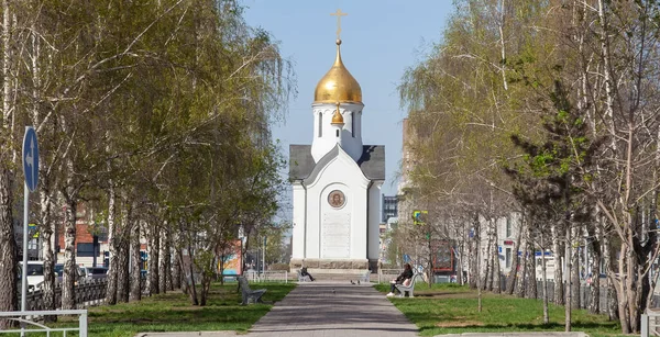 Chapel named after St. Nicholas the Wonderworker in Russian city of Novosibirsk is white with a golden dome next to a bench in an alley of birches located between two roads with different directions.