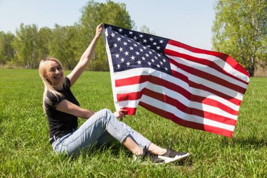 Siyah tişörtlü, kot pantolonlu ve spor ayakkabılı bir Rus kız yeşil çimlerin üzerinde oturuyor ve elinde huş ağacının arka planında rüzgarda sallanan Amerika Birleşik Devletleri bayrağını tutuyor..