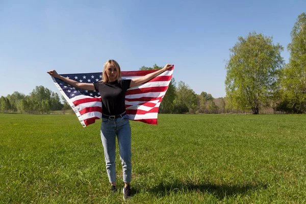 Siyah tişörtlü ve mavi kotlu bir kız arkasında Amerikan bayrağı olan bir Rus tarlasında duruyor, yeşil bir tarlanın ve ağaçların arkasında elinde tutuyor..