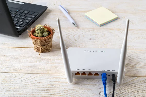 Beyaz Wi-Fi kablosuz yönlendirici, beyaz ahşap bir masada dizüstü bilgisayarın yanında. İnternet kablosu takılı Wlan yönlendirici bir ev ya da ofisteki masaya takılı. Arka görünüm.