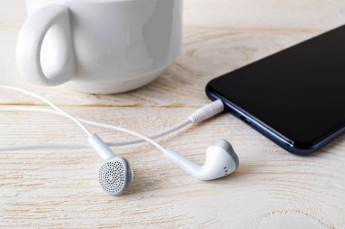 Beyaz kulaklıklar ve akıllı telefon beyaz ahşap bir masada fincanın yanında. Müzik ve iletişim için dokunmatik ekranlı modern kişisel cihaz. Ön görünüm.