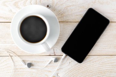 Akıllı telefon, beyaz kulaklıklar ve beyaz ahşap bir masada bir fincan sıcak kahve. Müzik ve iletişim için dokunmatik ekranlı modern kişisel cihaz. Üst görünüm.