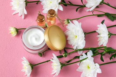 Altın kapaklı açık cam bir kavanozda yüz kremi, iki şişe kasımpatı esaslı yağ ve pembe arka planda beyaz kasımpatı. Güzellik, cilt bakımı ve kozmetoloji konsepti. 