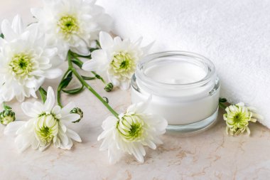 Açık cam bir kavanozda yüz kremi ve beyaz havlu yanında beyaz kasımpatı çiçekleri. Spa, güzellik, cilt bakımı ve kozmetoloji konsepti. Ön görünüm.