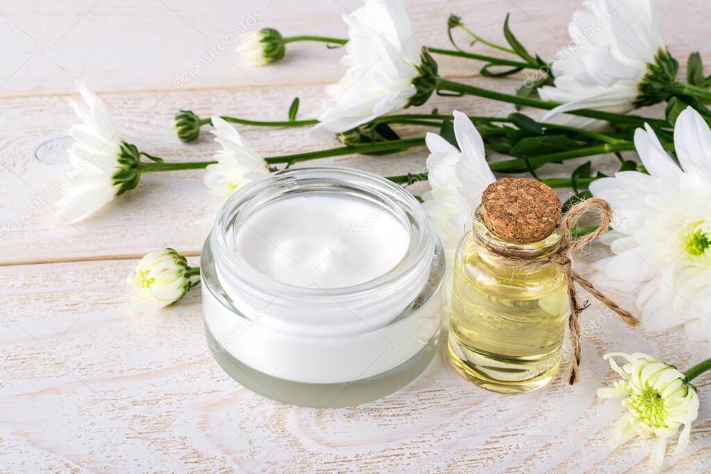 Facial cream in an open glass jar, chrysanthemum essential oil in a glass bottle and white chrysanthemums on a wooden surface. Spa, beauty, skincare and cosmetology concept. 