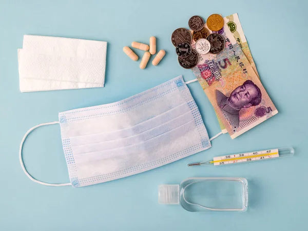 Disposable surgical mask, pills, thermometer, hand sanitizer and chinese yuan on a blue background. Medical mask reduces the risk of infection. Airborne disease protection concept.