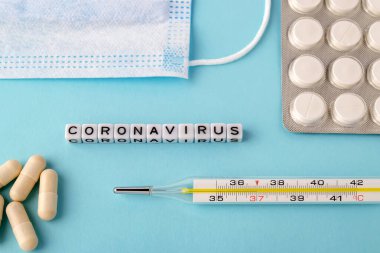 Kelime Coronavirus, cerrahi maske, termometre ve mavi arka planda haplar. Tıbbi maske enfeksiyon riskini azaltır. Havadan bulaşan hastalıkları koruma kavramı.
