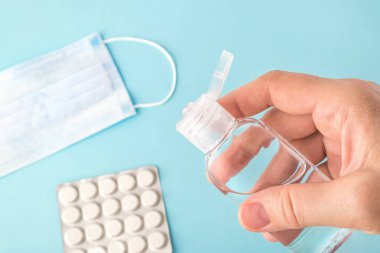 Kadın eli, ameliyat maskesi ve haplarla mavi bir yüzey üzerinde antibakteriyel el el jeli tutuyor. Enfeksiyon riskini ve hava yoluyla bulaşan hastalıkları koruma kavramını azaltmak.