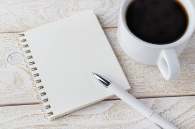 Açılmış bir yay defteri, beyaz tükenmez kalem ve beyaz ahşap desenli bir yüzey üzerinde bir fincan kahve. İş, eğitim, günlük tutmak ve konsept notlar yazmak. Boşluğu kopyala.