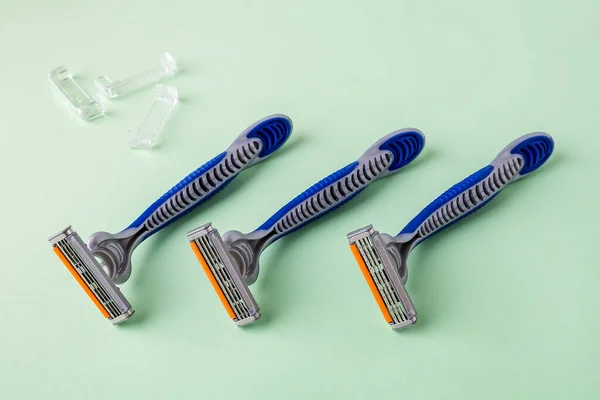 Three new disposable shaving razors with triple blade lie in a row on a pastel green background. Silver blue plastic razor with orange lubricat strip. Personal hygiene accessory for man and woman.