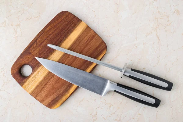Professional chef knife and a sharpening steel on a wooden cutting board over a kitchen table. Modern kitchen utensils made of high carbon molybdenum vanadium steel. Top view.