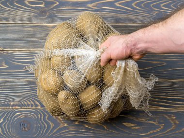 Erkek eli, PP ağ torbasını 10 kg çiftlik patatesiyle kaldırıyor. Polipropilen ağ çuvalı ve koyu ahşap zeminde 10 kilo organik patates. Toptan taze sebze almak için. Üst görünüm.