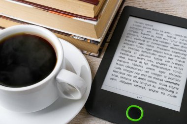 E-kitap okuyucusunun yakın çekimi, bir fincan sıcak kahve ve bir yığın sıradan kağıt kitap. Boşluğu e-kitap ekranına kopyala. Zevk ve eğitim için e-okuma. Ön görünüm.