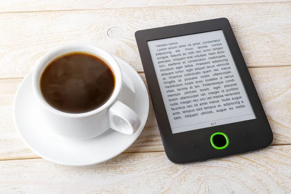 Beyaz ahşap bir masada sıcak kahve fincanının yanındaki e-kitap okuyucusuna uzayı kopyala. Zevk ve eğitim için e-okuma. Ön görünüm.