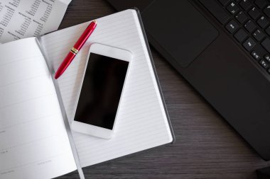 Yansıtıcı ekranlı akıllı telefonu kapat ve ofis masasına kağıt defter koy. Arka planda dizüstü bilgisayarlı bir ofis. Metin için alanı kopyala