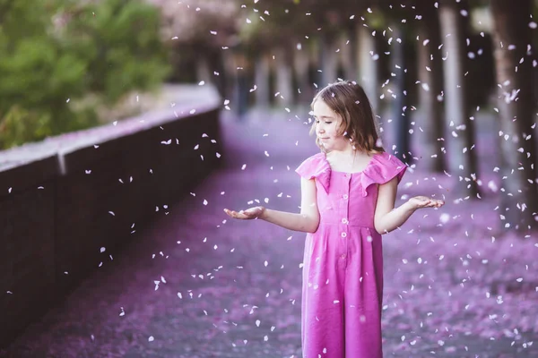 Hermosa chica en vestido rosa en el parque de flores de cerezo en un día de primavera, pétalos de flores que caen del árbol — Foto de Stock