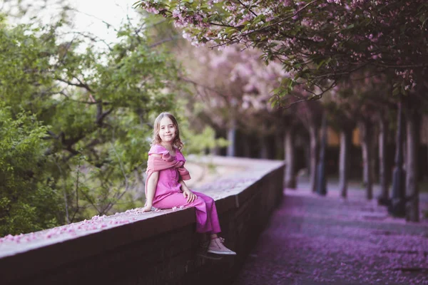 Beautiful girl 10 y.o. in pink dress sit in cherry blossom park on a spring day, flower petals falling from the tree — Stockfoto