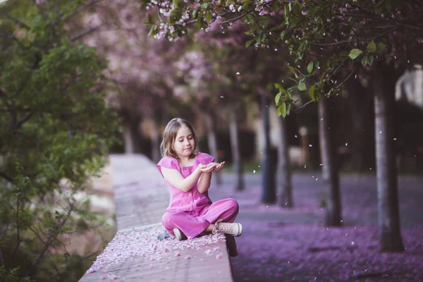 Beautiful girl 10 y.o. in pink dress sit in cherry blossom park on a spring day, flower petals falling from the tree — Φωτογραφία Αρχείου