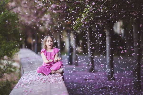 Beautiful girl 10 y.o. in pink dress sit in cherry blossom park on a spring day, flower petals falling from the tree — Stockfoto